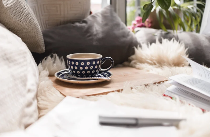 Kop koffie op een plank op de bank naast kussens met een boek en pen ernaast